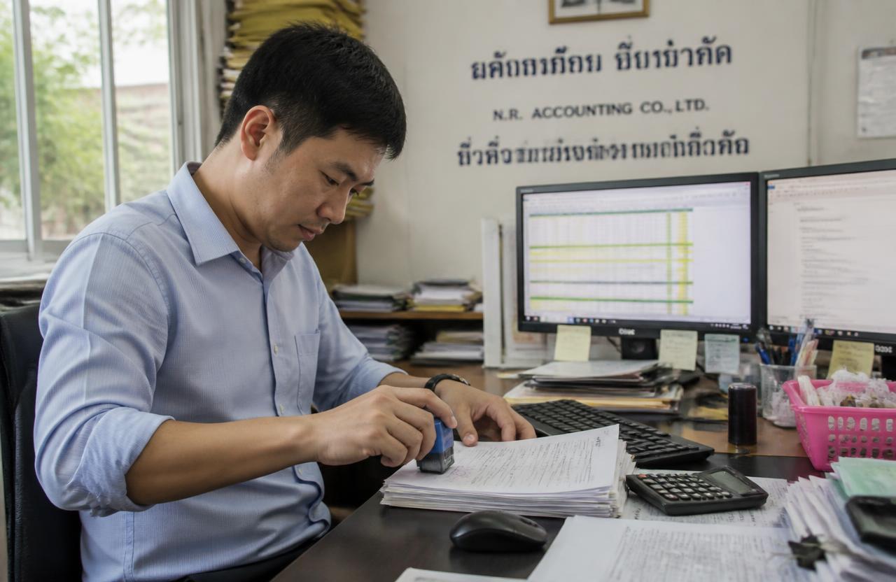 พนักงานบัญชีประทับตราและจัดเรียงหนังสือรับรองการหักภาษี ณ ที่จ่ายบนโต๊ะทำงานในสำนักงานไทย